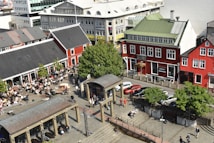 A lively urban scene featuring a busy plaza surrounded by buildings with distinct architectural styles. People are seated outdoors at cafes, enjoying the sunny weather. Red and white buildings with large windows line the street, and several cars are parked along the adjacent road. Leafy green trees add a touch of nature to the area.