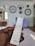 A hand holding a white Samsung TV remote with multiple buttons including Netflix and Prime Video. The background displays a living room setting with bicycle wheels mounted on a wall as decoration and a shelf with blue lighting. Natural light enters through a window, illuminating the room.
