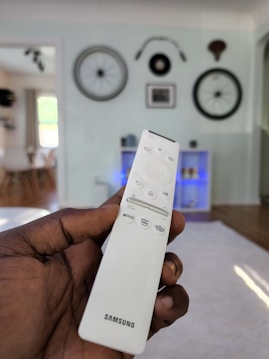 M3U PLAYLIST, A hand holding a white Samsung TV remote with multiple buttons including Netflix and Prime Video. The background displays a living room setting with bicycle wheels mounted on a wall as decoration and a shelf with blue lighting. Natural light enters through a window, illuminating the room.