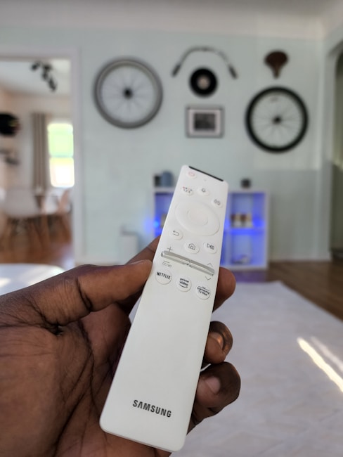 A hand holding a white Samsung TV remote with multiple buttons including Netflix and Prime Video. The background displays a living room setting with bicycle wheels mounted on a wall as decoration and a shelf with blue lighting. Natural light enters through a window, illuminating the room.