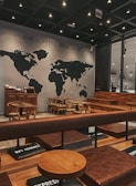 A cozy coffee shop interior featuring a world map mural with the phrase 'OUR COFFEES COME FROM' above it. The seating consists of wooden chairs and tables, with padded benches in a warm brown leather tone. The ambiance is modern with soft, dim lighting from numerous small ceiling lights, creating an inviting atmosphere.