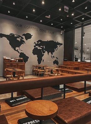 A cozy coffee shop interior featuring a world map mural with the phrase 'OUR COFFEES COME FROM' above it. The seating consists of wooden chairs and tables, with padded benches in a warm brown leather tone. The ambiance is modern with soft, dim lighting from numerous small ceiling lights, creating an inviting atmosphere.