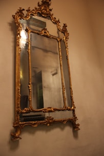 An ornate, vintage mirror with a gilded, decorative frame featuring detailed carvings and a reflective surface. It is mounted on a plain, beige wall.