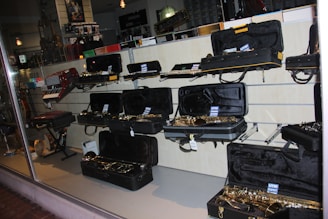 A musical instrument store display featuring an array of instruments, primarily saxophones, in open black cases on shelves. Additional instruments such as a keyboard accordion are visible. The lighting creates a reflective atmosphere on the glass window.