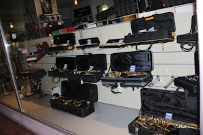 A musical instrument store display featuring an array of instruments, primarily saxophones, in open black cases on shelves. Additional instruments such as a keyboard accordion are visible. The lighting creates a reflective atmosphere on the glass window.