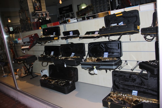 A musical instrument store display featuring an array of instruments, primarily saxophones, in open black cases on shelves. Additional instruments such as a keyboard accordion are visible. The lighting creates a reflective atmosphere on the glass window.