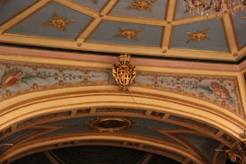 An ornate ceiling with intricate floral designs and golden embellishments on a light blue background. The architecture features detailed molding and a prominent emblem with a crown at the center of an archway. The edges showcase decorative floral patterns with pastel colors.