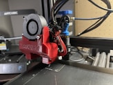 A close-up view of a 3D printer's extruder assembly, featuring a red component labeled BLOKINHEAD. Various wires and a small fan are connected to the extruder, which is part of a larger 3D printer setup visible in the background. The workspace has a modern and technical appearance with equipment and tools.