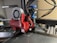A close-up view of a 3D printer's extruder assembly, featuring a red component labeled BLOKINHEAD. Various wires and a small fan are connected to the extruder, which is part of a larger 3D printer setup visible in the background. The workspace has a modern and technical appearance with equipment and tools.
