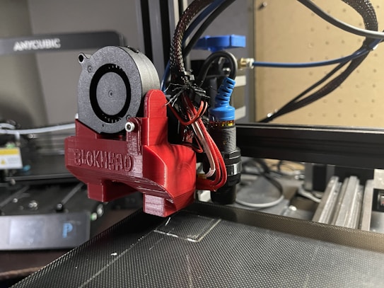 A close-up view of a 3D printer's extruder assembly, featuring a red component labeled BLOKINHEAD. Various wires and a small fan are connected to the extruder, which is part of a larger 3D printer setup visible in the background. The workspace has a modern and technical appearance with equipment and tools.
