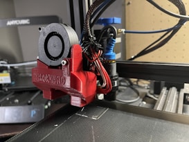 A close-up view of a 3D printer's extruder assembly, featuring a red component labeled BLOKINHEAD. Various wires and a small fan are connected to the extruder, which is part of a larger 3D printer setup visible in the background. The workspace has a modern and technical appearance with equipment and tools.