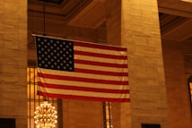 A large American flag hangs inside an ornate building with a classic architectural style. The setting features tall stone columns and a detailed ceiling, illuminated by warm lighting and a grand chandelier.