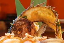 A close-up view of a beautifully plated dish featuring a whole fish presented in a curved position, garnished with various herbs. The background includes green leaves and some blurry elements that indicate a dining setup.