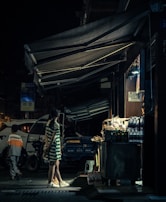 A nighttime street scene featuring a young person in a striped dress standing near a small, well-lit street vendor stall. Various products like bottles and snacks are displayed. Another person in the background walks away, while a bicycle and vehicle add to the urban setting.