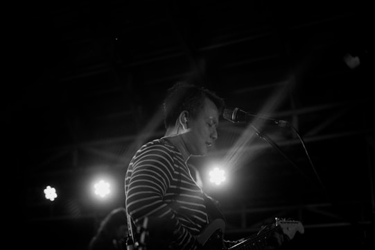 A musician is performing on stage, captured in black and white. Wearing a striped shirt, the person is playing an electric guitar while singing into a microphone. Bright stage lights illuminate in the background, creating a dramatic and focused atmosphere.