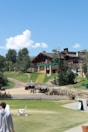 A rustic wooden lodge surrounded by lush greenery and a bright blue sky. There are several pathways and chairs on grass in the foreground. A few people are walking or lounging around the area.