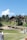 A rustic wooden lodge surrounded by lush greenery and a bright blue sky. There are several pathways and chairs on grass in the foreground. A few people are walking or lounging around the area.