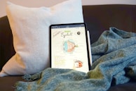 A tablet displaying an educational diagram of a human eyeball resting on a dark sofa with a light cushion behind it. A blue-green textured blanket is draped around the tablet, and a white stylus is placed beside it.
