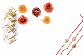 The image features a traditional arrangement for a festive occasion. On the left, there is a stack of square sweets with silver leaf on top, known as kaju katli. In the center are three orange and yellow marigold flowers arranged around a small, intricate clay oil lamp. To the right are three decorative rakhis, featuring beads and traditional designs, tied with red threads.