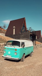 A turquoise and white vintage Volkswagen van is parked on a gravel driveway in front of a modern A-frame house with a large wooden deck and a chimney. The house is set on a hillside with a stone retaining wall and minimal vegetation.