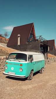 A turquoise and white vintage Volkswagen van is parked on a gravel driveway in front of a modern A-frame house with a large wooden deck and a chimney. The house is set on a hillside with a stone retaining wall and minimal vegetation.