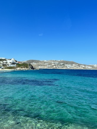 A clear, vibrant seascape with turquoise waters in the foreground transitioning to darker blue as it meets the horizon. Small white buildings dot the rocky coastline under a brilliant blue sky. The hills behind are sparse with vegetation.