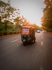 An auto rickshaw is speeding down a road lined with trees during a sunset. The motion blur creates a sense of movement. The sun casts a warm glow over the scene, adding drama and vibrancy.