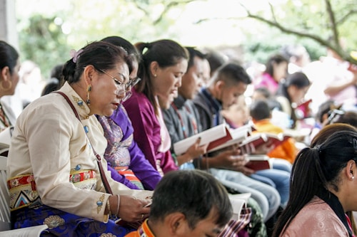 A group of people sitting closely together, each holding and reading from books. They appear to be focused and engaged, dressed in colorful, traditional clothing. The scene suggests a cultural or community gathering, possibly a workshop or seminar.