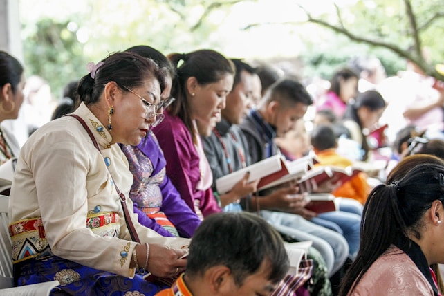 A group of people sitting closely together, each holding and reading from books. They appear to be focused and engaged, dressed in colorful, traditional clothing. The scene suggests a cultural or community gathering, possibly a workshop or seminar.