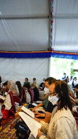 A group of people sit closely together on the ground inside a tent, wearing masks and reading from documents. The setting is casual and communal, with a colorful fabric border lining the upper part of the tent interior. Natural light filters in from an opening in the tent, and there is a patterned carpet on the ground.