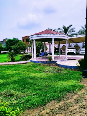 A well-manicured garden features a circular gazebo with a red-tiled roof supported by white columns. It is surrounded by lush green grass and complemented by palm trees in the background. There is outdoor seating under the gazebo, and nearby a covered area with parked cars is visible. The sky above is partly cloudy.