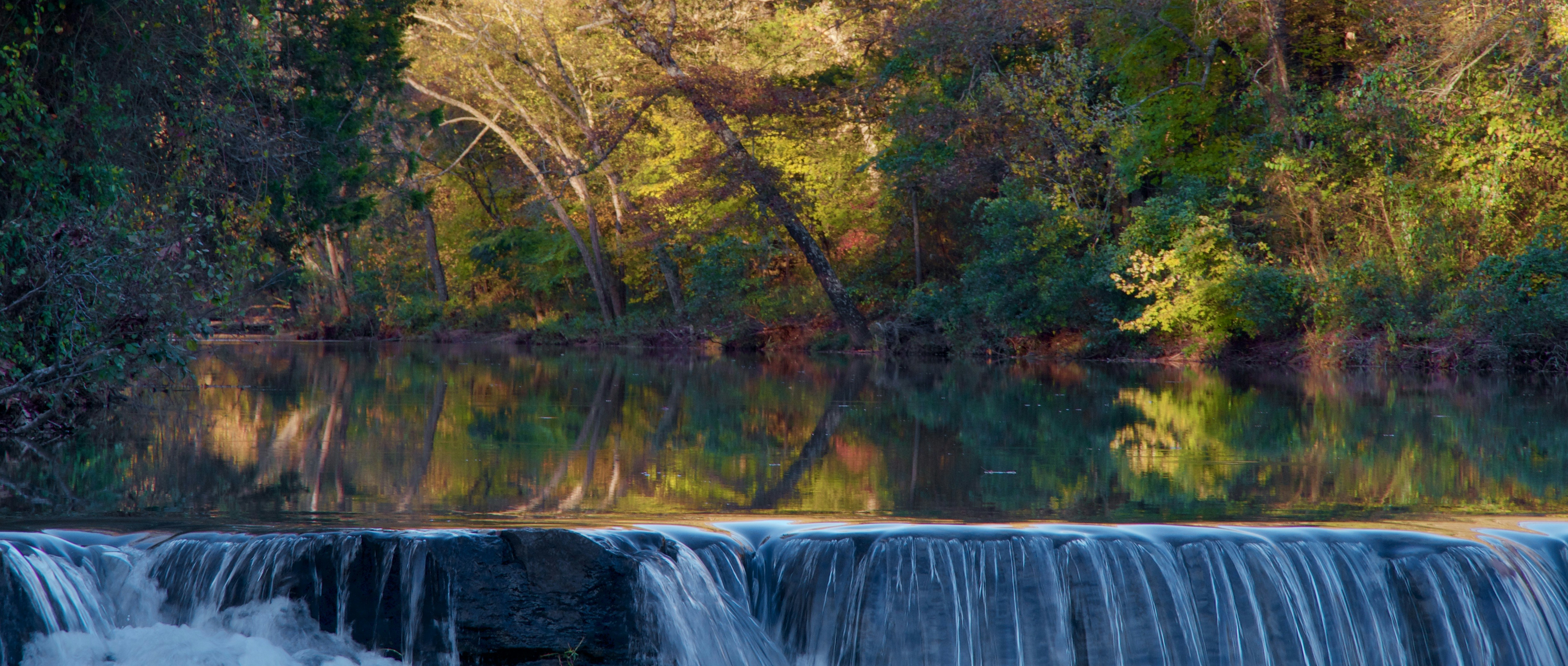 Waterfall cascading over a rocky ledge into a forested pool surrounded by colorful autumn foliage.