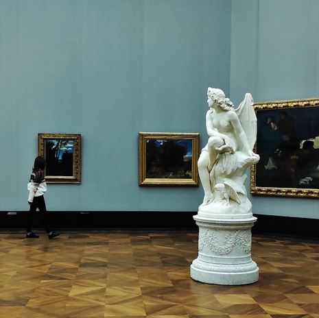 A large white marble statue of a winged figure seated on a pedestal is prominently displayed in an art gallery. The sculpture is positioned near several framed paintings hung on the room's walls. A person, appearing to be a visitor, is walking past the art pieces, looking towards them. The gallery's interior features a wooden floor and light blue walls.