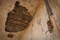 A damaged ceiling with a large section of plaster missing, exposing dark structural beams. The surrounding walls are old and peeling, showing signs of severe wear and decay. On one side, there is a rusted pipe extending vertically, with corrosion visible on the metallic parts.
