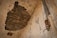A damaged ceiling with a large section of plaster missing, exposing dark structural beams. The surrounding walls are old and peeling, showing signs of severe wear and decay. On one side, there is a rusted pipe extending vertically, with corrosion visible on the metallic parts.