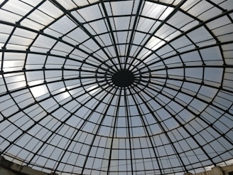 A large glass dome structure with a geometric grid pattern, radiating from a central point in a circular fashion. The dome is composed of glass panels framed by metal bars, creating a symmetrical and architectural appearance.