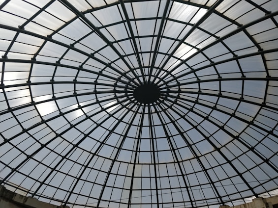 A large glass dome structure with a geometric grid pattern, radiating from a central point in a circular fashion. The dome is composed of glass panels framed by metal bars, creating a symmetrical and architectural appearance.