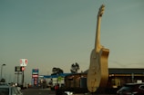 A large, golden guitar sculpture is prominently displayed in front of a building, with a recognizable fast food sign and a gas station in the background. Cars are parked nearby under a clear sky.