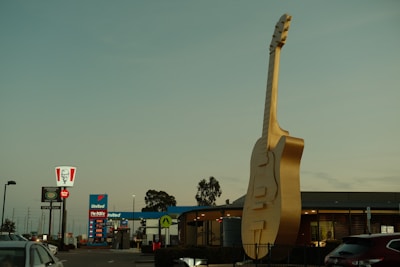 A large, golden guitar sculpture is prominently displayed in front of a building, with a recognizable fast food sign and a gas station in the background. Cars are parked nearby under a clear sky.