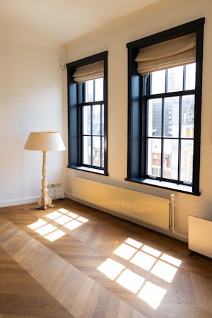 Two tall windows with black frames and beige roman shades allow natural light to flood a room with light-colored walls. Below the windows, a white radiator is mounted on the wall. A wooden floor with a geometric pattern reflects the sunlight. A beige table lamp with a wood-carved base is positioned next to the left window.