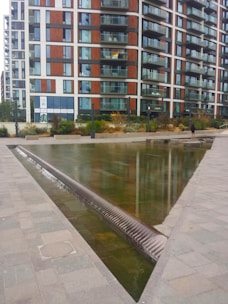A modern apartment building with a combination of glass and wooden paneling, featuring numerous balconies. In front of the building, a shallow triangular water feature with a smooth stone surface is visible, bordered by paved walkways. A person stands near the edge of the water feature, adding a sense of scale to the image. The surrounding area includes some landscaping with shrubs and low plants.