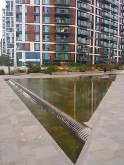A modern apartment building with a combination of glass and wooden paneling, featuring numerous balconies. In front of the building, a shallow triangular water feature with a smooth stone surface is visible, bordered by paved walkways. A person stands near the edge of the water feature, adding a sense of scale to the image. The surrounding area includes some landscaping with shrubs and low plants.