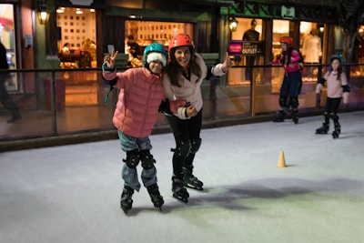 Two people wearing helmets and protective gear are ice skating indoors. They seem joyful, with one person making a peace sign and both smiling at the camera. In the background, other skaters can be seen, and there is a shopfront with warm lighting.