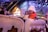 A group of children are enjoying a ride inside an amusement park decorated with winter-themed elements. One child wearing a white hat and orange jacket appears to be looking towards the camera with a smile. The background features festive lights, snowflakes, and structures designed to look like small houses with snowy roofs.