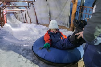 A child dressed in winter clothing sits in a blue inflatable snow tube at the base of an icy slide. A person assists by handling the child’s boot. The area is surrounded by snow and wooden beams, with protective sheets along the slide.