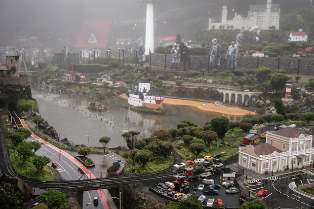 A detailed miniature model depicts a small town with roads, cars, trees, and buildings. There are tiny representations of people wearing raincoats, a bridge spanning a river, and a lighthouse near the bank. In the background, there are castle-like structures on a hill, and everything is set under a misty sky.