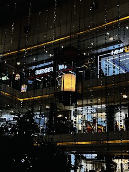 A multi-level shopping mall interior with various illuminated store signs, including those for a burger place and fashion brands. Decorative vertical hanging elements create an artistic effect. Warm ambient lighting complements the overall modern design.