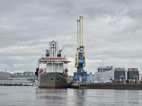 A large ship is docked at a port with a prominent white and black structure, featuring multiple antennas and satellite dishes on its upper deck. A massive yellow and blue crane towers beside the ship, indicating an industrial harbor environment. The background includes buildings and additional smaller ships, under a cloudy sky.