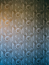 An Art Deco style pattern features intricate geometric shapes and swirling lines, arranged in a symmetrical and repetitive design. The pattern uses a gradient of blue and gold hues, creating a stylish and elegant visual effect.