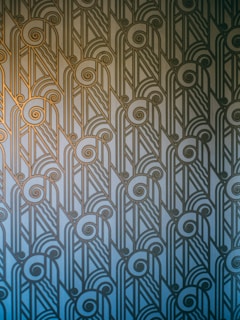 An Art Deco style pattern features intricate geometric shapes and swirling lines, arranged in a symmetrical and repetitive design. The pattern uses a gradient of blue and gold hues, creating a stylish and elegant visual effect.